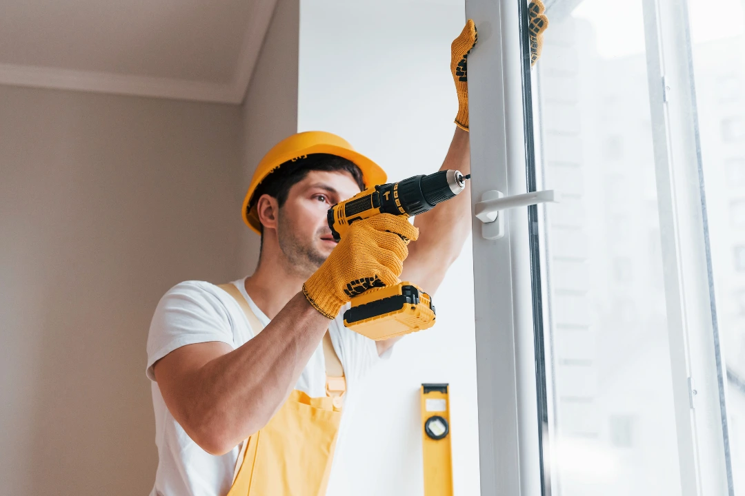 A construction worker in a yellow hard hat and gloves uses a power drill to install a window